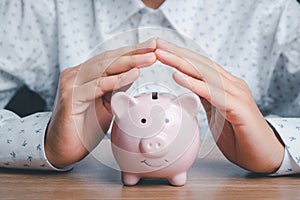 Piggy bank under hands in gesture of protection on wooden desk