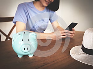 Piggy bank on wooden table with blurry background of woman using smart phone, booking hotel online, saving money for vacation