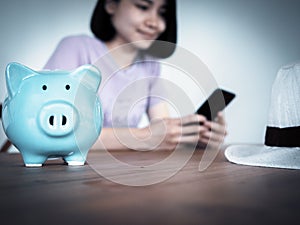 Piggy bank on wooden table with blurry background of woman using smart phone, booking hotel online, saving money for vacation