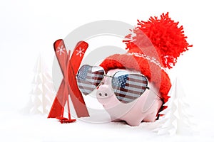 Piggy box with red hat with pompom and sunglasses shape heart with USA flag standing next to red ski and ski sticks on snow and ar