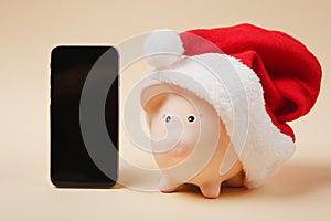 Piggy money bank with Christmas hat, mobile phone with blank empty screen on beige background. Money