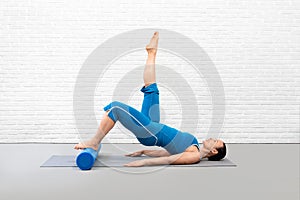 Pilates with foam roller. Adult caucasian woman in blue sportswear does gluteal bridge with one leg up, in fitness