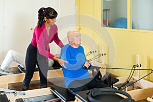 Pilates trainer helping elderly man do hands stretch on the machine in gym