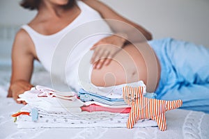 Pile of baby clothes, stuff and pregnant woman in home interior