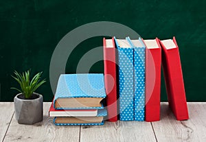 Pile of books in colorful covers and plant in pot on wooden table with green blackboard background. Distance home education. Back