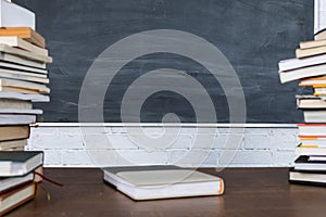 A pile of books stacked on either side of a school desk. One book is in the center of the table top. There is a clean