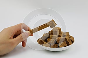 A pile of brown sugar cubes on a white plate woman hand hold wooden tweezers on white background