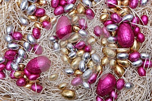 Pile of colorful pale pastel foil wrapped chocolate easter eggs in pink,  silver and gold, on pale shreaded cream nest.