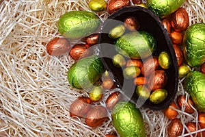 Pile of colourful foil wrapped chocolate easter eggs in green, orange, yellow and gold with two halves of a large dark eggs.