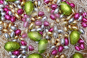 Pile of colourful pastel foil wrapped chocolate easter eggs in green, pink, yellow, silver and gold, on pale shreaded cream nest.
