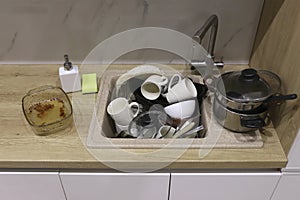 Pile of dirty dishes like plates, cups pot and cutlery in the white kitchen in the light beige granite sink