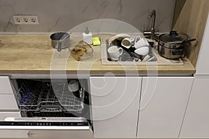Pile of dirty dishes like plates, cups pot and cutlery in the white kitchen in the light beige granite sink. Open