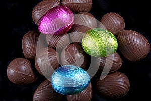 Pile of foil wrapped & unwrapped chocolate easter eggs in pink, blue & lime green, against a black background