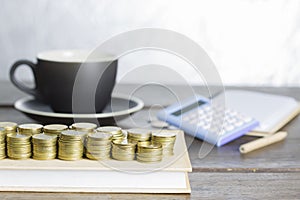 Pile of gold coin on cover book and black coffee cup blue calculator on wooden table.