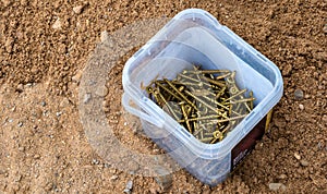 A pile of golden wood screws in a plastic container, laying in the dirt