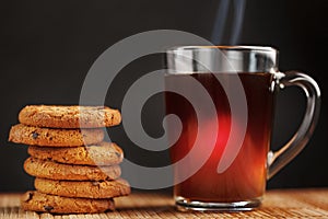 A pile of oatmeal cookies with chocolate chips and a mug of fragrant black hot tea in on a bamboo substrate, on a dark background