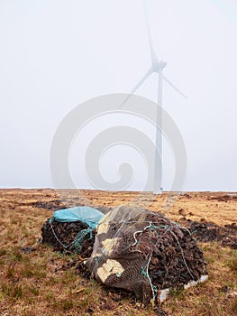 Pile of peat fossil fuel in foreground, wind power turbine in a fog in the background. Old and new source of energy. Ecology