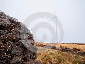 Pile of peat fossil fuel in foreground, wind power turbine in a fog in the background. Old and new source of energy. Ecology