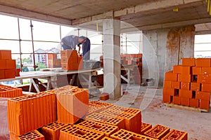 Pile of red blocks, workers stand on wood scaffold and build wall with bricks and mortar, building site