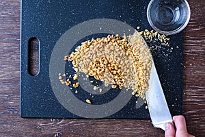 Pile of rough chopped and fine chopped walnuts on a black cutting board, womanÃ¢â¬â¢s hand on chef knife, glass bowl.