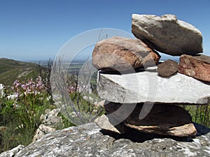 A pile of stacked rocks overlooking a view from a mountain top,  indigenous vegetation blurred in the background