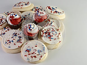 Pile of sugar cookies covered in white frosting with red, white and blue sprinkles and mini chocolate cupcakes with red and white