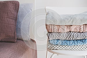 Pile of warm sweaters on white chair standing in bedroom near neat and tidy bed