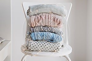 Pile of warm woolen sweaters wrapped in decorative Christmas lights on white chair