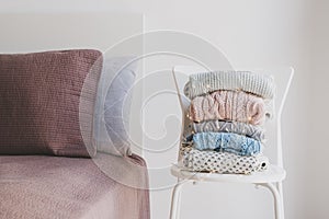 Pile of warm woolen sweaters wrapped in decorative lights on white chair and clean and neat bed