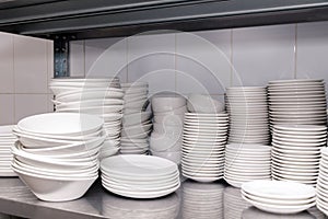 A pile of white clean ceramic porcelain plates on a metal rack in the back of the restaurant. Concept of preparation for banquet,