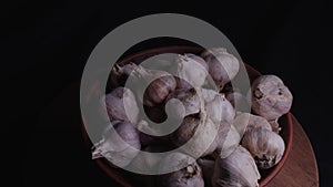 Pile of whole bulbs of garlic in ceramic bowl on table. Harvest of unpeeled vegetables on black background.