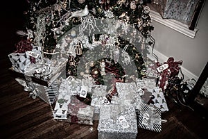 Pile of wrapped Christmas presents with name tags and bows in pile on wooden floor under old fashioned Christmas tree with