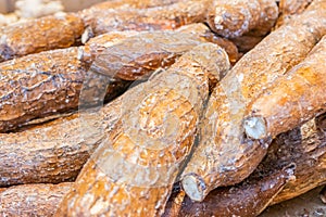 Pile of yuca root tubers under bright light, with dark brown skin, being sold at a farmer`s market. Also called cassava root.