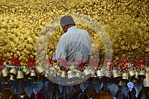 Pilgrim pasting gold foils onto golden rock at the Kyaiktiyo Pagoda, Myanmar with row of small bells in foreground