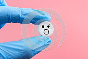 A pill with a screaming emoticon in a hand in a medical glove on a pink background. Painkiller concept