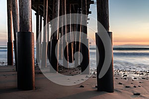 Pillars of Ventura Pier at sunset. Beach sand and rocks; smooth ocean and colored sky.