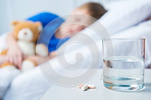 Pills and glass of water on table and boy lying in hospital bed with teddy bear