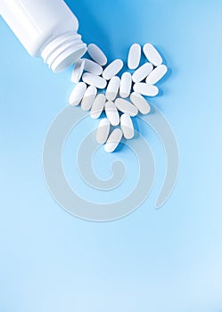 Pills of vitamin in the shape of heart with the opened white plastic container on soft blue background
