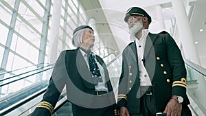 Pilots, escalator and people at airport for travel, talking and conversation in low angle. Mature, captain and first