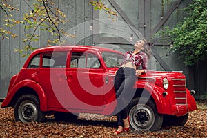 Pin-up girl in jeans and a plaid shirt is leaning on a russian red retro car.