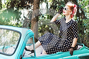 Pin-up girl wearing vintage clothes, sitting on old car hood.