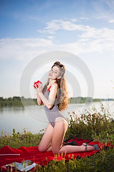 Pin up style portrait of a girl in striped swimsuit with long curly hair and red lipstick holding a ball on river bank in summer