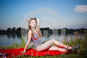 Pin up style portrait of a girl in striped swimsuit with long curly hair and red lipstick sitting on river bank at summer day.