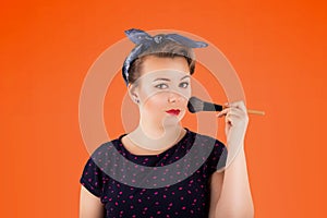 Pin-up woman portrait with bright colors and beautiful make-up in front of an orange background holding a make-up br in her hands.