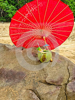 Pina colada tropical beverage drink sitting on a rock at a beach under a red umbrella