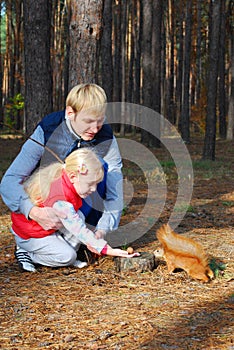 In the pine forest father and daughter feed a squirrel nuts.