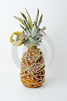 Pineapple decorated like a Christmas tree. Gold beads garland. Golden shining reflective disco ball. White background