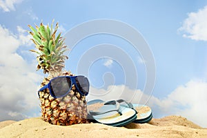 pineapple fruit with sunglasses and flip flop sandals on the beach sand against a blue sky with clouds, summer vacation