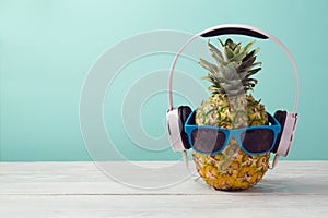 Pineapple with headphones and sunglasses on wooden table over mint background. Tropical summer vacation and beach party.