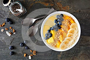 Pineapple, mango smoothie bowl with coconut, bananas, blueberries and granola, above view table scene on slate
